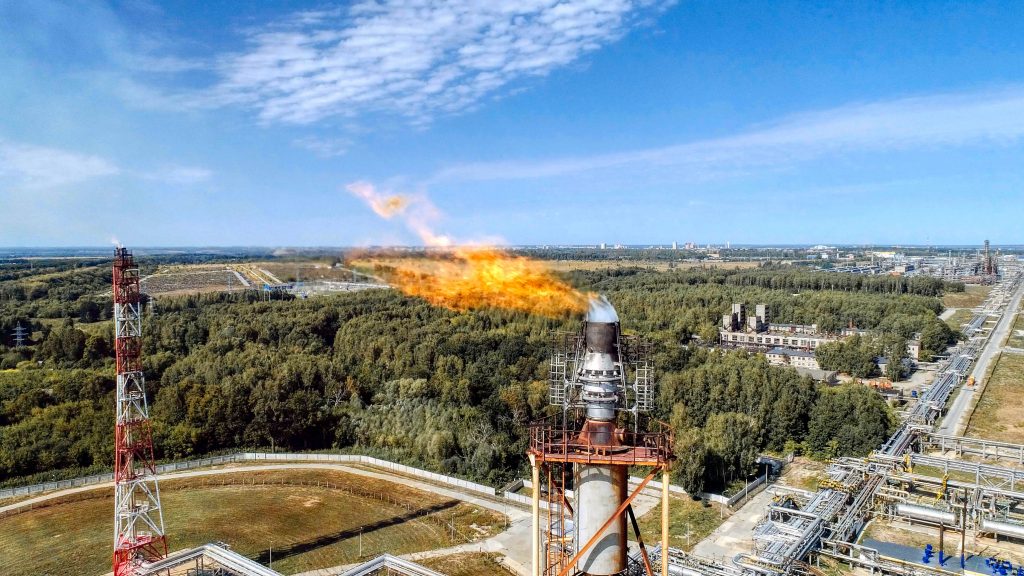 Torchère brûlant des gaz à effet de serre dans une zone industrielle, visible en fond de paysage verdoyant.